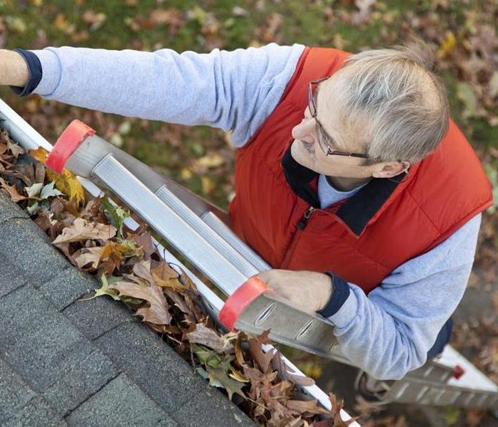 A man on a ladder cleaning leaves out of the gutter on his house